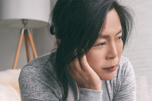 A middle-aged asian woman sitting on her couch, resting her head on her hand, suffering from adrenal fatigue. Get treatment for adrenal fatigue syndrome from Tanya Zboril, NP of TBT Medical in Regina.