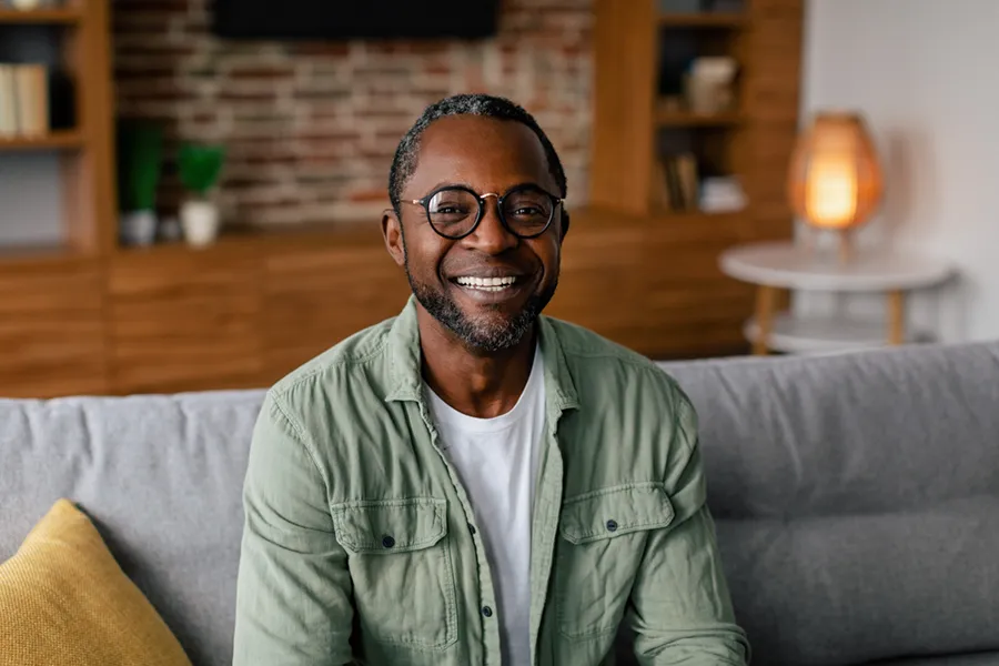 Andropause-Doctor A middle aged black man in a green button up shirt sits on his couch smiling, happy with his treatment for andropause and low testosterone from Tanya Zboril, NP of TBT Medical in Regina.