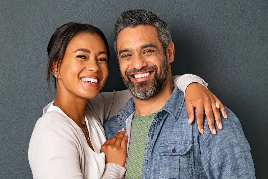 A middle-aged couple with their arms around each other standing against a gray background and smiling, happy with the bioidentical hormone therapy they received from Tanya Zboril, NP of TBT Medical in Regina.