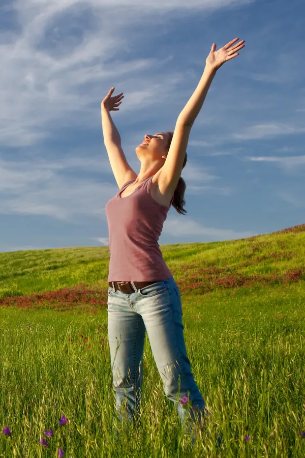 A woman in a tank top and jeans looks up with her arms outstretched to the sky in a field after successful treatment for Grave's Disease from Tanya Zboril, NP of TBT Medical in Regina.