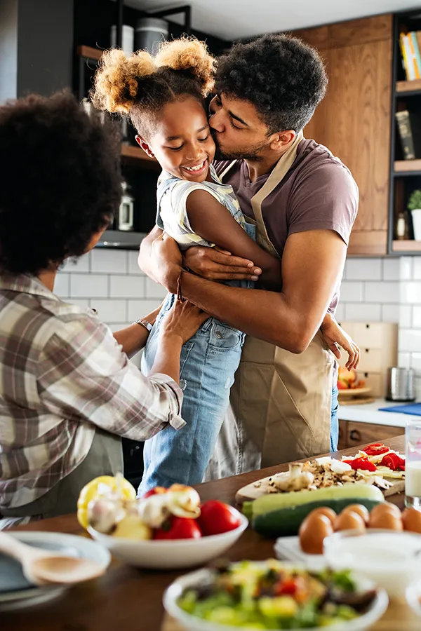 A man holds his daughter and kisses her cheek as the family makes a healthy dinner in the kitchen. Get help for digestive issues and gut health from Tanya Zboril, NP of TBT Medical in Regina.