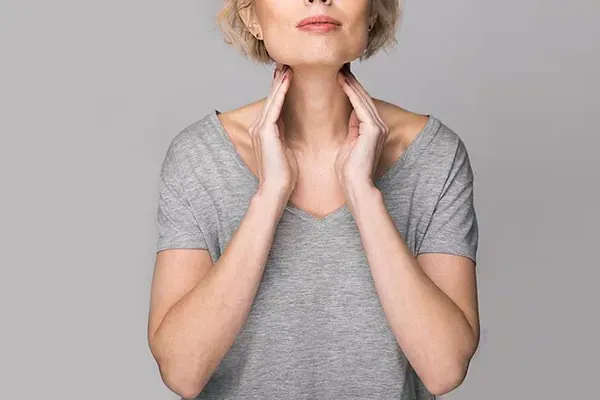 An older blonde woman in a gray t-shirt holds her hands to her neck checking for signs of thyroid problems. Get help for Hashimoto's Thyroiditis from Tanya Zboril, NP of TBT Medical in Regina.