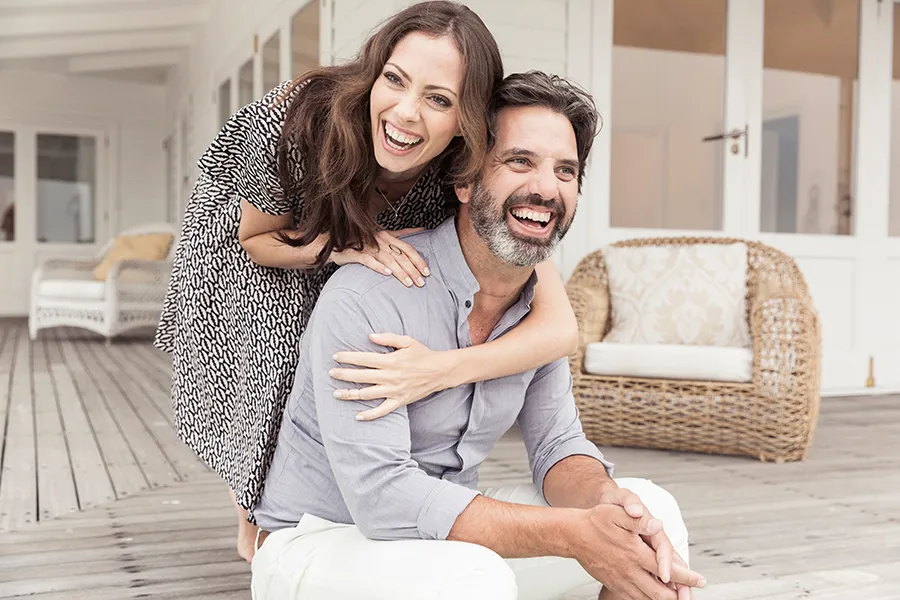 A middle-aged couple on a wooden porch with wicker furniture; the woman is playfully hugging the man from behind. They are benefiting from the health and wellness services from Tanya Zboril, NP of TBT Medical in Regina.