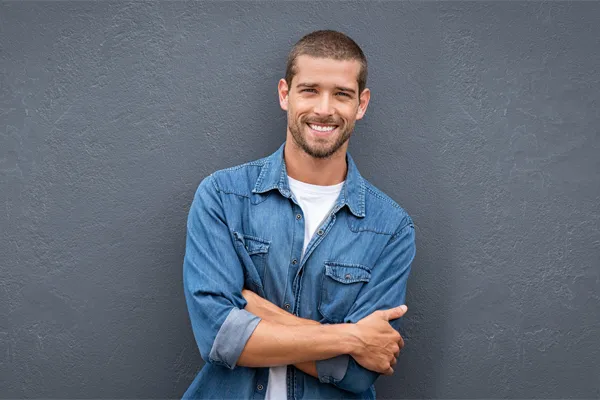 A man in a denim shirt stands smiling against a gray-blue wall, pleased with his testosterone hormone therapy from Tanya Zboril, NP of TBT Medical in Regina.