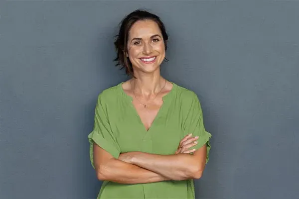 A woman in a green shirt stands smiling against a gray-blue wall, pleased with her hormone therapy from Tanya Zboril, NP of TBT Medical in Regina.