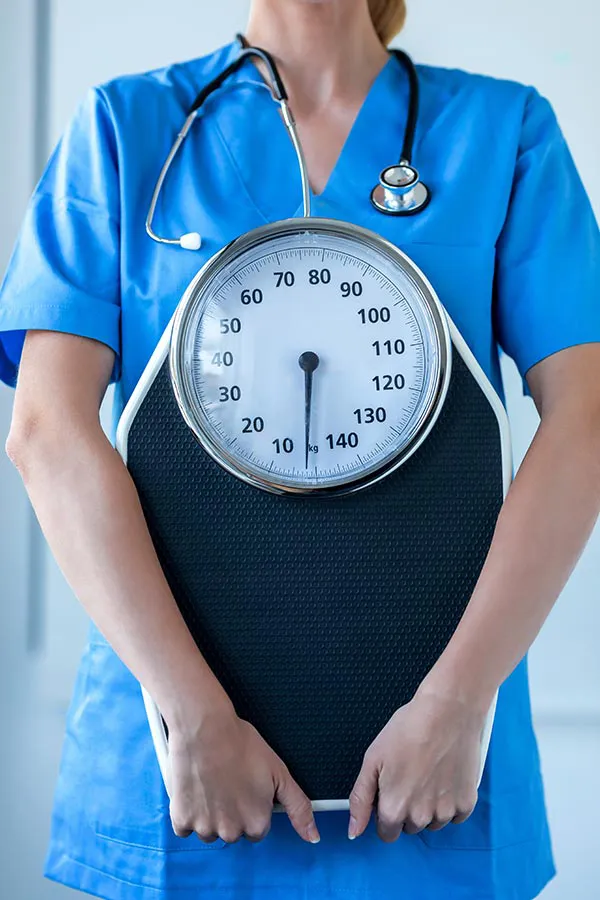 A nurse in blue scrubs with a stethoscope hanging around her neck holds a scale, representing weight loss treatments from Tanya Zboril, NP of TBT Medical in Regina.