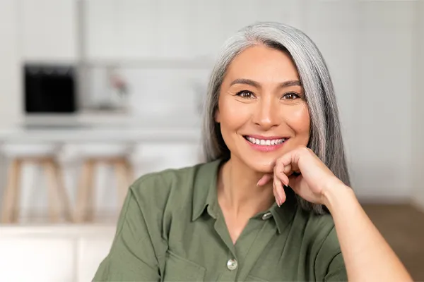 Menopause-Clinic An asian woman with long gray hair and a green shirt sitting and smiling on her couch, happy with her menopause care from [NAME} in Regina.