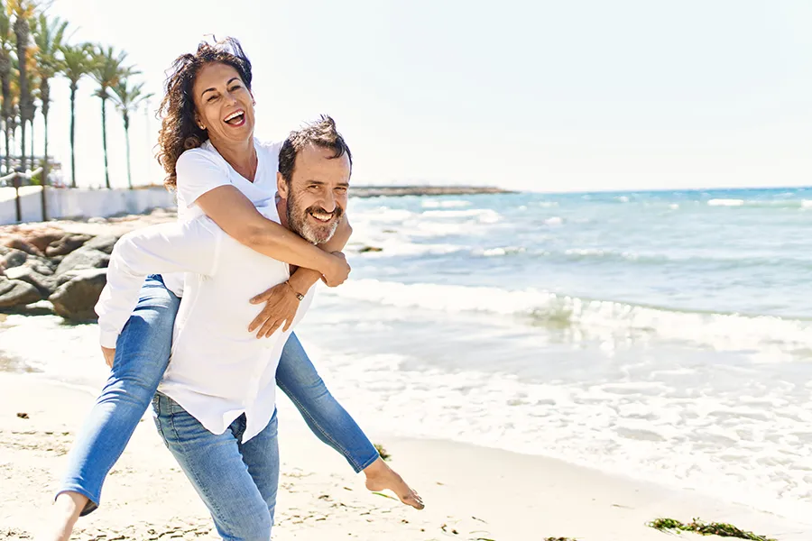 A woman piggybacks on her husband as the middle-aged couple have fun at the beach. Get NAD Therapy from Tanya Zboril, NP of TBT Medical in Regina.