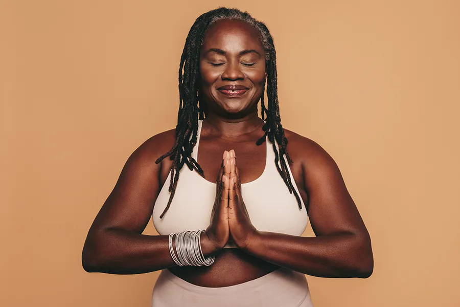 Obesity-Clinic An older African American woman confidently posing against a warm background smiling with hands in a prayer pose. Tanya Zboril, NP of TBT Medical treats obesity using functional medicine in Regina.