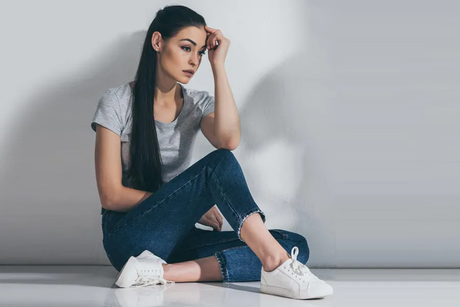 A young brunette woman in a gray t-shirt and jeans sits against a gray wall with her head resting on her hand and a distressed expression. Get help for Polycystic Ovarian Syndrome (PCOS) from Tanya Zboril, NP of TBT Medical in Regina.