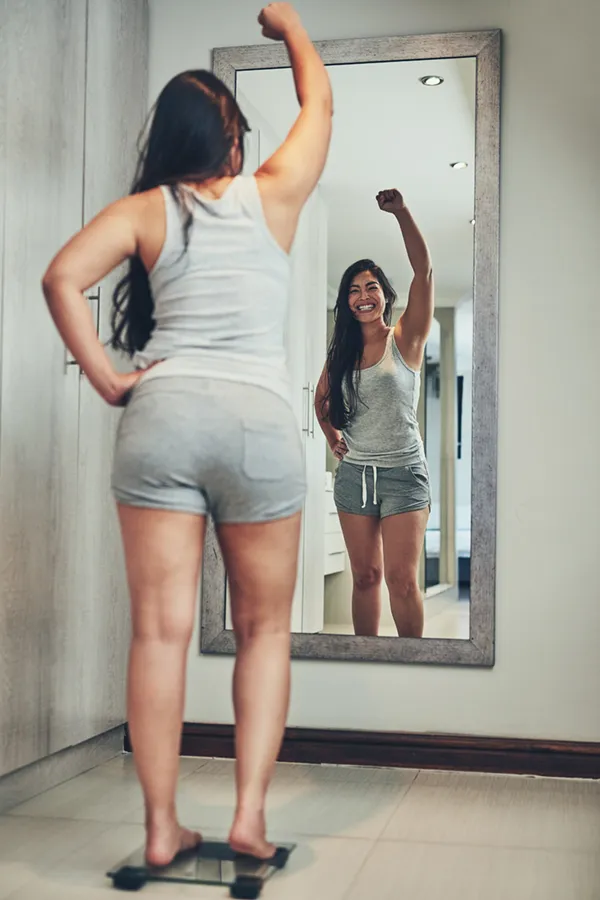 A woman in workout clothes stands on a scale looking in the mirror and pumping her hands in celebration of her successful Tirzepatide treatment from Tanya Zboril, NP of TBT Medical in Regina.