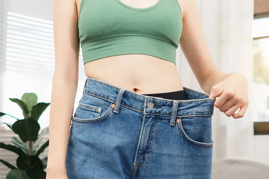 A close-up of a young woman in a green crop top, holding out her oversized blue jeans to show significant weight loss. Zepbound Weight Loss Injections offered by Tanya Zboril, NP of TBT Medical in Regina.